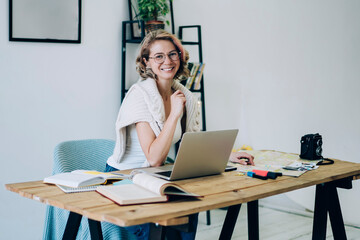 Portrait of cheerful female freelancer in trendy wear working remotely in cozy home interior with blank pictures advertising, happy hipster girl sitting at desktop with modern laptop computer