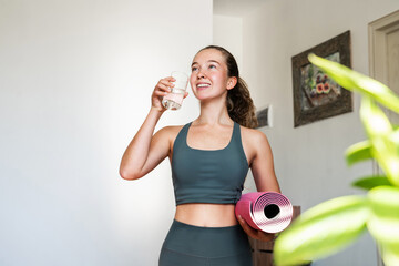 Sporty cheerful young woman drinking water after training at home, fit girl in green sportswear standing at home with rubber mat in hands, concept of healthy lifestyle, fitness and hydration