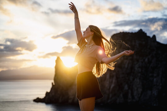 Young woman dancing at sunset on a summer day among sea and mountains background, stunning golden sunbeams shine on the playful beautiful girl twirling, feeling of freedom at sunset, travel holidays