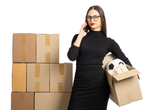 Woman in a black dress holding a cardboard box with a soccer ball, talking on the phone, standing next to a stack of boxes. Perfect for business, logistics, or retail themes. - Powered by Adobe