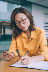 Thoughtful woman writing notes while browsing phone