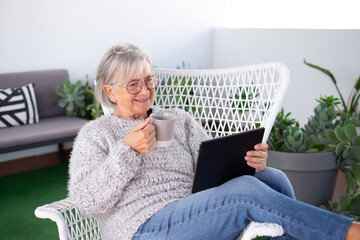 Attractive senior gray haired woman holding a coffee cup relaxing at the armchair in the balcony at home looking at digital tablet