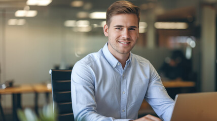 Fototapeta premium A young Caucasian man in a light blue shirt smiles confidently while working at a desk in a modern office environment.