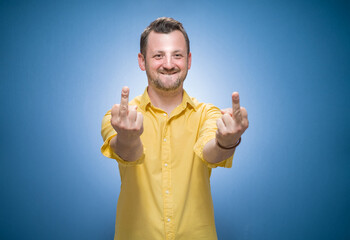 Young man showing fuck off gesture over blue background, dresses in yellow shirt. Male person with obscene gesture giving the middle finger and looking to camera