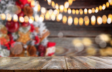 Christmas background. Empty wooden table on foreground with garland of Christmas lights and blurred christmas tree in background.