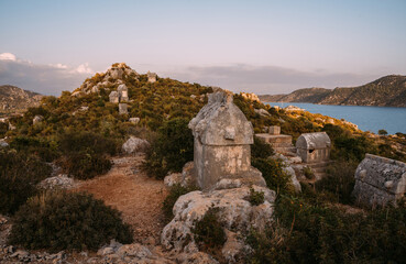 Fototapeta premium Stone sarcophagus from ancient Simena Kaleköy city in Turkey. Mediterranean sea surrounded by rugged hills listed morning light. Archaeological treasure of Lycia.