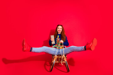 Fototapeta premium Full body photo of attractive young woman ride sledge spread legs gift dressed blue christmas clothes theme party on red color background