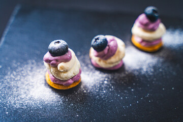 Fototapeta premium The cake lost its shape next to the two well-standing. Mini desserts on a black mica board with blueberry cream and blueberries, sprinkled with powdered sugar