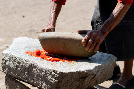Manos de una mujer campesina moliendo aji rojo en un batan al aire libre.