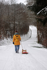 Young woman in yellow jacket pulling a wooden sled on a snowy path by a road. Tenager girl on a winter walk in forest with snow covered trees, Outdoor stroll on a quiet snowy day.