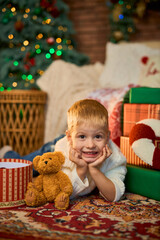 Happy boy of 3 years old lies on floor of house next to Christmas tree with side of garlands and gift boxes at home. Christmas and New Year time traditions.