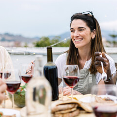 Happy young woman enjoying summer dinner celebration at home terrace gathered with group of friends, drinking red wine and enjoying barbecue food