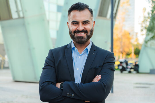 Confident businessman with arms crossed posing outdoors, wearing a suit and smiling, representing success and professionalism, potential Turkish or Indian heritage in a modern urban business setting
