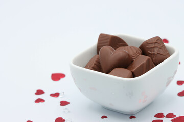 White Dish Full Of Chocolates Surrounded By Red Heart Confetti And Confetti Gems On White Background