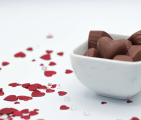 White Dish Full Of Chocolates Surrounded By Red Heart Confetti And Confetti Gems On White Background