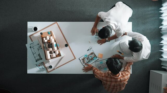 Group of skilled designer looking at model while discussing about building. Top view of architect engineer explain idea while talking and writing blue print scattering at meeting table. Alimentation.