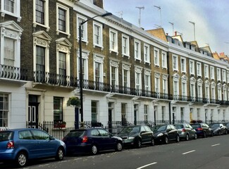 Naklejka premium A row of traditional English townhouses with intricate architectural details, alongside parked cars on a quiet street. The image showcases a classic urban residential scene.