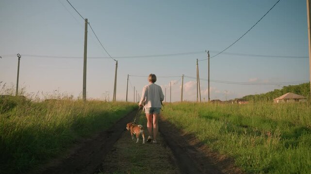 Back view of woman walking with dog on dirt path lined with electric poles, holding leash while both stroll through grassy rural field under bright sunlight, distant buildings visible in background