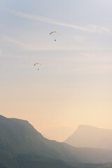 Two paragliders in the air fly over the mountains at summer sunrise.