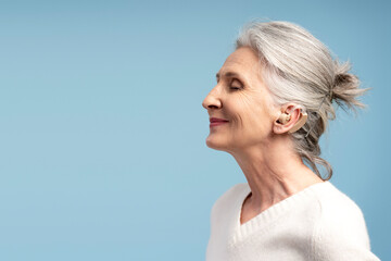 Smiling, happy senior woman having hearing aid, with closed eyes, posing isolated on blue background
