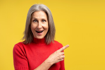 Excited, smiling senior woman with open mouth pointing finger on copy space, standing