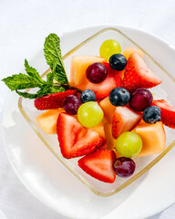 Fruit salad in glass bowl on white table with strawberries, grapes, blueberries, melon, and mint leaves