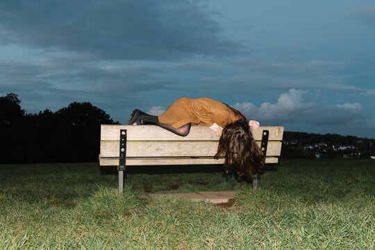 Woman Lying On Park Bench Under Cloudy Sky At Dusk