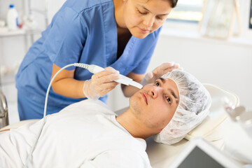 Fototapeta premium Female cosmetic doctor deftly wields Skin Scrubber attachment, carefully treating gentleman's skin during procedure.