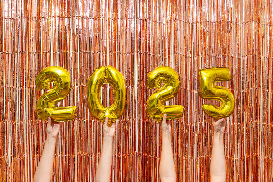 Hands holding gold 2025 balloons with tinsel backdrop

