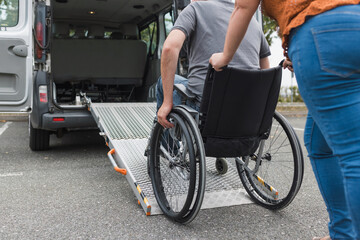 Female assistant pushing a man with disability, a wheelchair user up the car ramp into the van....