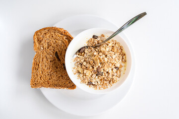 Plate with simple breakfast centered in the frame. Bowl with muesli and yogurt and a slice of brown bread next to it. White soft background with natural light.