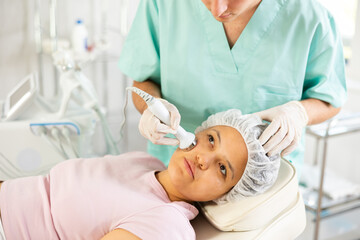 Naklejka premium Young male cosmetologist performs facial ultrasound procedure with machine to young female client