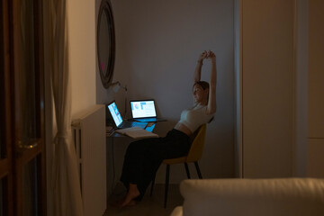 Woman stretching at her desk while working late on laptop