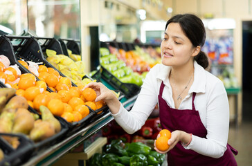In vegetable shop,Asian woman seller in apron selects ripe sweet tangerines for regular customer in window.Local farm products