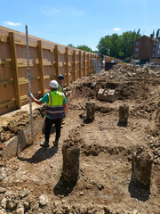 Construction site with two workers in safety gear inspecting excavation area with soil piles, wooden retaining wall, and foundation piles. Outdoor site under sunny sky.