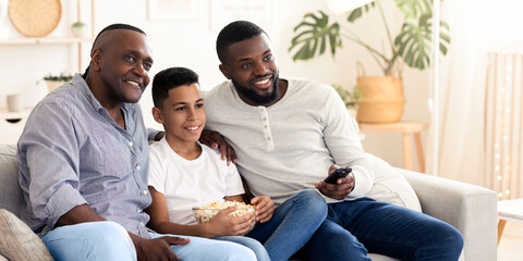 Cheerful african grandpa, father and son enjoying comedy show on tv, relaxing together on couch in living room, eating popcorn