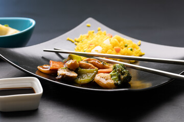 Metal chopsticks placed on a plate serving traditional Chinese Hunan chicken with vegetables, served alongside fried rice, wonton soup, and soy sauce.