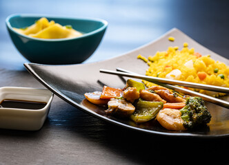 A traditional Chinese dish, Hunan chicken, served on a black square plate with fried rice. Accompanied by wonton dumpling soup, a small bowl of soy sauce, and metal chopsticks crossed on the plate.