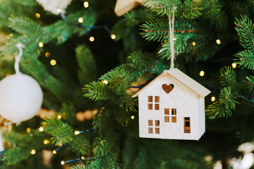 Handcrafted wooden house ornament with heart shaped cutout hanging on Christmas tree branch with warm lights background, creating festive atmosphere of winter holidays. Wallpaper, backdrop