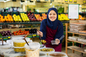 Young woman seller in hijb and apron sells green olives in grocery store