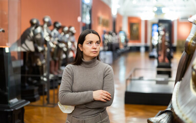 Portrait of interested adult female tourist visiting exhibition of medieval armor in armory of historical museum ..