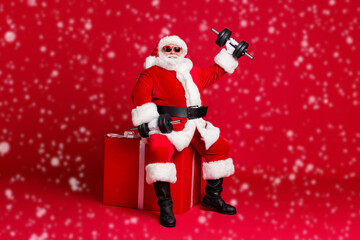 Full length photo of retired old man white beard hold dumbbell sit large box show kid grandpa still strong wear santa x-mas costume sunglass headwear boot isolated red color background