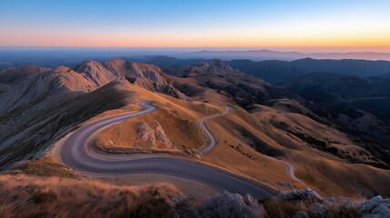 Fototapeta premium Winding mountain road seen from above, surrounded by rugged landscape under a clear sky at sunrise or sunset, with mountainous terrain and distant horizon