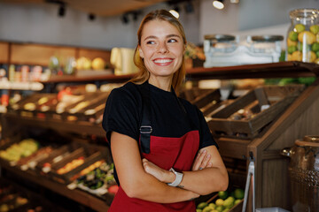 A cheerful woman in a grocery store stands proudly, showcasing fresh produce and creating a warm, welcoming atmosphere