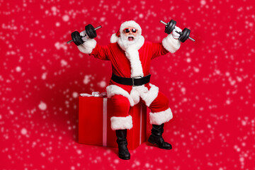 Full length photo of retired old man white beard sit giftbox hold dumbbells crazy strength scream wear x-mas santa costume glove coat belt sunglass cap boot isolated red color background