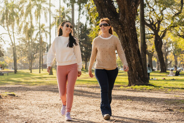 Two women walking through a city park, smiling and chatting in the afternoon sunlight.