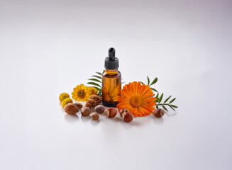 A small amber glass bottle with a dropper sits on a white surface next to yellow and orange flowers, green leaves, and brown nuts.