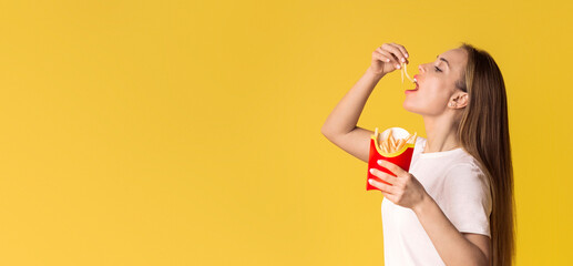 Cheat Meal Day Concept. Hungry Millennial Lady Eating French Fries Serving, Standing Over Yellow Studio Background, Side View With Free Space