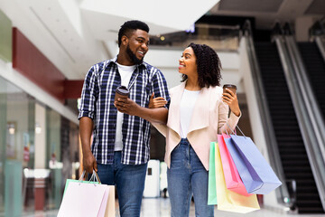 Shopping Lovers. Cheerful Black Couple Walking With Takeaway Coffee And Shopper Bags In Mall, Enjoying Seasonal Sales And Discounts, Happy African Spouses Doing Purchases Together, Free Space