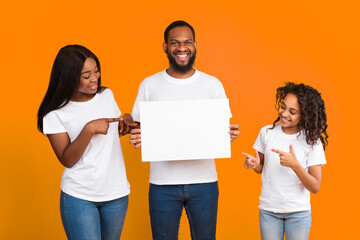 Place For Your Ad. Portrait of smiling black man holding empty blank board, family pointing fingers at square paper for mockup template, free space for text or design isolated on yellow studio wall